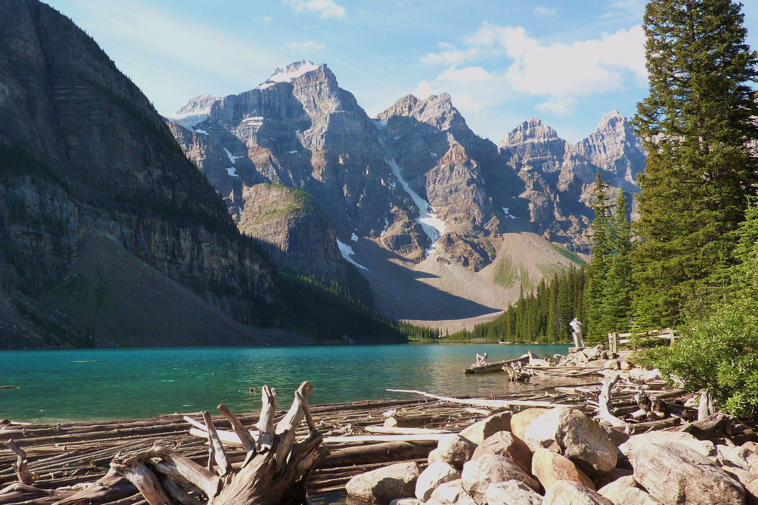 A lake with a backdrop of mountains, with pine trees lining the right side, and scree slopes on the left